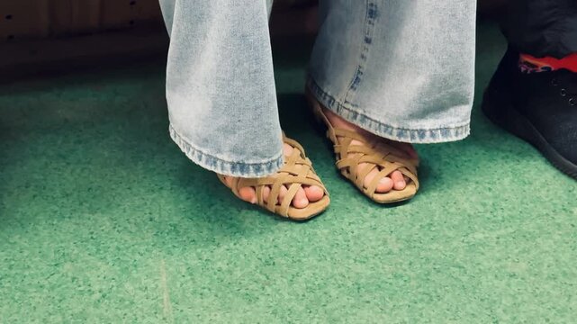 Close Up Feet In Woven Sandals Flared Denim Hem Grazing Braided Leather Straps On Green Carpet, Shopper Trying On Shoes During Boutique Fitting, Warm Retro Mood, Summer Styling Detail.