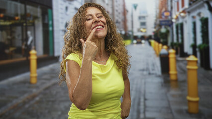Fototapeta premium Woman points finger to teeth while smiling broadly in a yellow top on a wet narrow cobblestone street; confidence celebration.