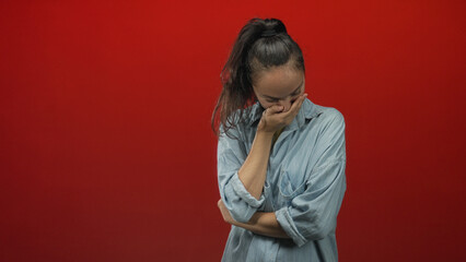 Fototapeta premium Woman with hand covering mouth, eyes closed and crossed arms, wearing denim shirt against red studio backdrop; sadness introspection.