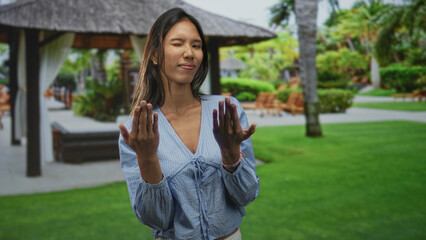 Fototapeta premium Thai woman smiles and holds hands forward showing fingernails and rings, wearing blue blouse in resort gazebo by palm trees; serenity self care.