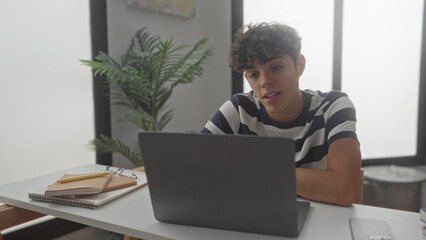 Teen boy typing on laptop at desk in building, fingers on keyboard and smiling while notebooks and phone nearby; concentration success.