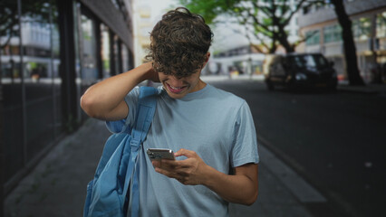 Teenage boy tugging shirt collar while holding smartphone and grimacing on a street; frustration...