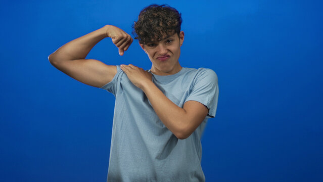 Teenage man flexes bicep and pulls sleeve to show muscle in studio, wearing blue tshirt and smiling; confidence youth energy.