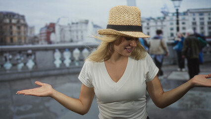 Fototapeta premium Woman with arms raised and open palms on city bridge wearing straw hat and white t shirt, hair blown by wind; playful wonder.
