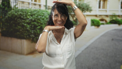 Fototapeta premium Senior hispanic woman frames face with hands, hand under chin and other over head while smiling in front of a building courtyard; joyful.