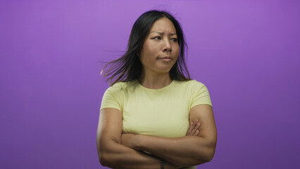 Fototapeta premium Young hispanic woman with crossed arms and furrowed brow standing in studio against vivid purple wall; annoyed defiance.