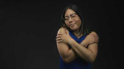 Fototapeta premium Woman wearing a blue dress and glasses hugging her shoulders with arms crossed in a black studio; self care acceptance serenity.