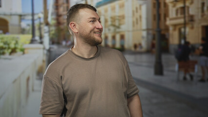 Obraz premium Man smiling and looking sideways on a city street in a short sleeved brown t shirt, beard visible; contentment curiosity travel.