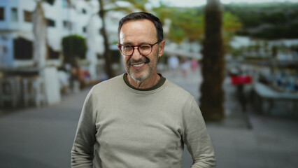 Mature hispanic man in urban park making quiet gesture with finger on lips, wearing glasses and sweater, surrounded by blurred city street scene.