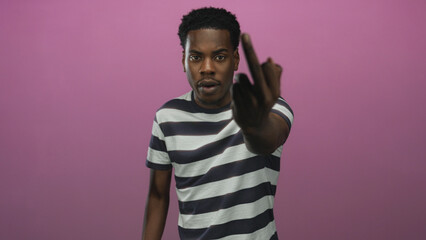 Obraz premium Young african american man in a striped shirt flipping middle finger in a pink studio; defiance rebellion.