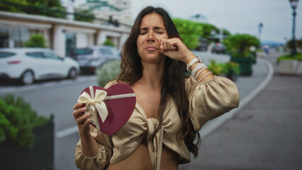 Fototapeta premium Woman holding heart shaped gift box wiping eye on city street with teary face and closed eyes; heartbreak sorrow.