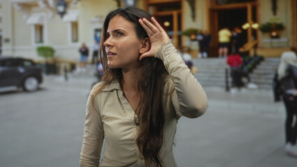 Fototapeta premium Woman cups hand to ear listening on a city street by building steps and crowd, profile visible; curiosity focus.