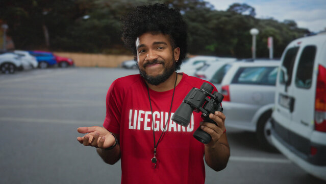 Man wearing lifeguard shirt holds binoculars on a busy city street with parked cars, showcasing an urban outdoor scene with a casual and relaxed vibe.