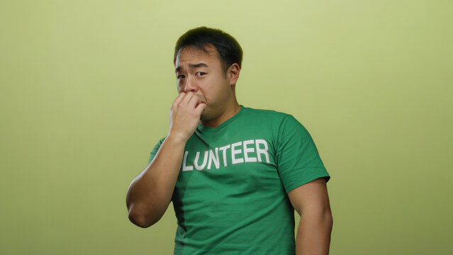 Young man wearing green volunteer shirt standing against yellow background with thoughtful expression, embodying introspection and engagement in isolated setting.