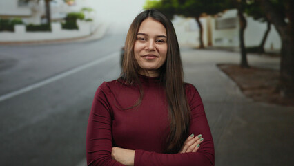 Fototapeta premium Woman smiling with arms crossed standing on an outdoor street, showcasing a confident and relaxed expression in a casual urban environment.
