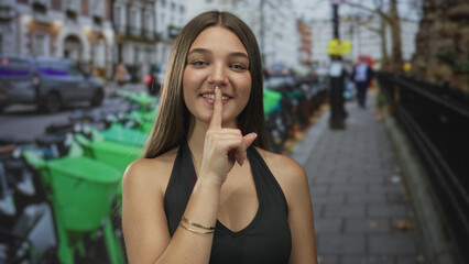Fototapeta premium Teenage girl finger to lips for silence beside green rental bikes parked on bustling city street; hush calm.