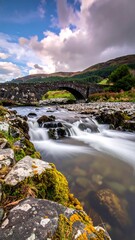 Old stone bridge over a flowing river, mountain backdrop, cloudy sky