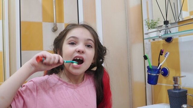 A happy little girl brushes her teeth in front of a bathroom mirror. Morning hygiene. A modern schoolgirl with red hair, the alpha generation. A child washes her face in the bathroom in the morning