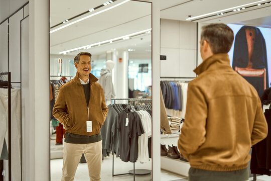 Man Trying On Brown Suede Jacket in Fashion Mall Store, Smiling At His Reflection In a Large Mirror