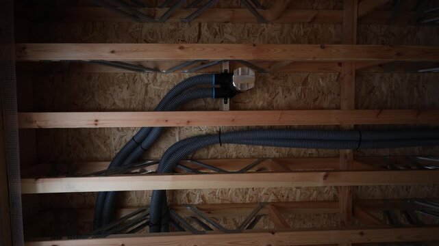 Slow pan shows exposed joists, OSB sheathing, wood fibre loose fill, grey corrugated ducts and manifold on stud, routed through truss webbing with cable ties in warm light.