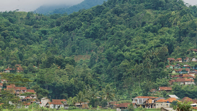 Residential houses nestled at the foot of a lush, forested mountain in Sumedang, Indonesia. Traditional village life in a tropical valley.