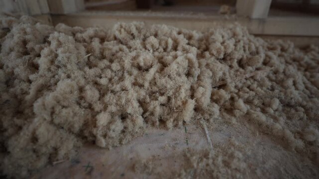 Close up pans along timber joists showing pale tan loose wood fibre filling floor voids, with green crosses on membrane and subtle dust motion during framing stage.