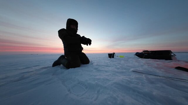 One person kneels on a frozen lake tending a line near a drill hole. A sled, bucket, and portable shelter sit nearby as pastel bands meet deep snow shadows.