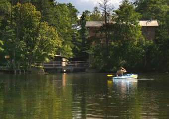 Obraz premium An unrecognizable person in a kayak seen from behind paddling on a beautiful and wild Krutynia river surrounded by dense green forest. Solitude and nature recreation in Poland.