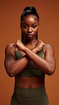Confident dark-skinned woman in an olive sports bra and leggings stands powerfully against a warm brown studio background, embodying fitness, strength