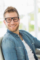 Adult male sitting smiling looking at camera by windows wearing black glasses denim shirt white tee