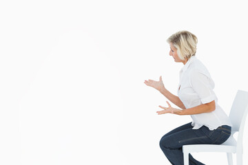 Senior woman is sitting on chair at right of backdrop, wearing white shirt, jeans, copy space