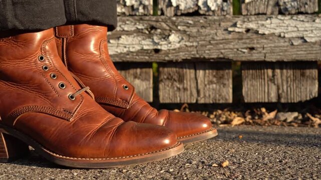 Two unlaced brown leather boots stand on gray pavement a weathered wooden fence in the background