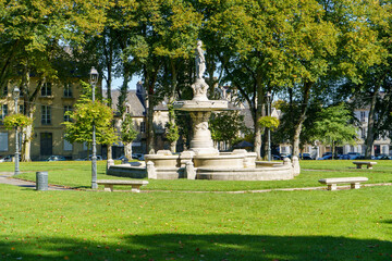 Obraz premium Majestic stone fountain in a sunny park, surrounded by green trees, scattered autumn leaves, and classic buildings.