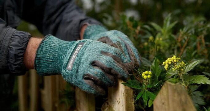 Gardener's hands in protective gloves holding wooden fence post near blooming plants