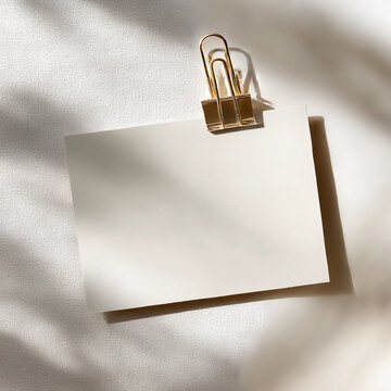 a blank white postcard, on the right side of it is a golden paper clip holding up two sheets of a4 paper together, shadows from window light on a textured cream background