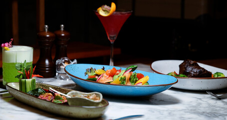 Gourmet dishes and vibrant cocktails sitting on marble table in upscale restaurant. Smoked salmon salad, braised meat, and craft drinks highlight luxury lifestyle and fine dining experience.