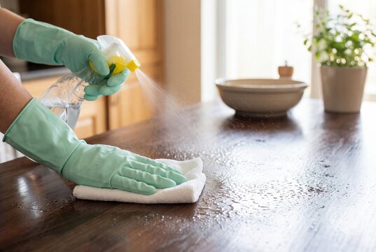 Hands wearing green gloves cleaning a wooden kitchen table with spray bottle and cloth
