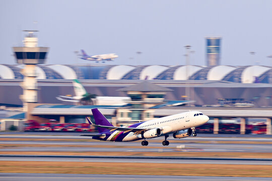 Passenger airplane takes off from busy airport runway, with terminal and control tower visible in background. scene captures dynamic motion of air travel