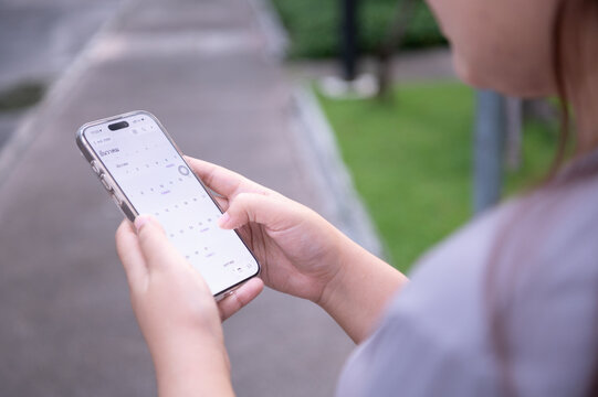 A person on a sidewalk holds a smartphone displaying a calendar app, with lush green grass and a pathway in the background.