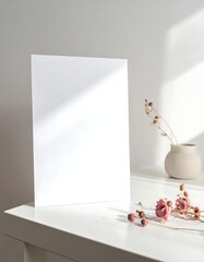 A stark white, rectangular card stands upright on a white surface, bathed in soft, natural light and shadows, with minimal adornments