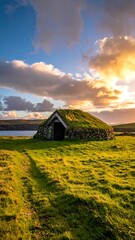 A stone hut with a grass roof sits on a grassy field with a lake and colorful sky at sunset