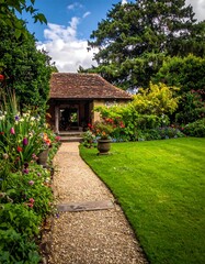 A stone path leads to a small brick building nestled amidst vibrant flowerbeds and lush greenery under a bright sky