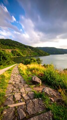 A stone path leads toward a tranquil lake under a dynamic sky. Lush greenery borders the pathway and hills surround the water