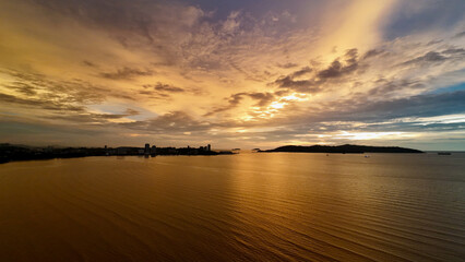 Golden sunset over tropical sea with city skyline on the horizon. Aerial view shows glowing water, dramatic clouds and urban coastline silhouetted against evening sky.