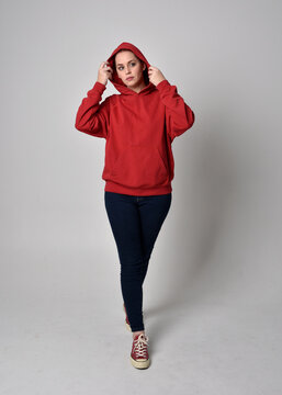 full length figure of female model wearing  basic contemporary casual clothing, red hoodie jumper, skinny jeans and sneakers. standing pose walking towards camera,  isolated on studio background.
