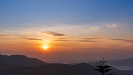 Fototapeta premium Sunrise over mountain layers with soft mist and tree silhouette creating a peaceful morning scene.