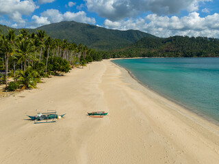 Fototapeta premium Aerial view of a wide sandy beach with two small traditional boats resting on the shore beside calm turquoise water. Palm trees line the coastline with forested hills and a quiet tropical bay in the b