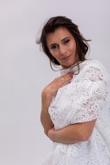 Woman posing in white lace garment with arm across body and subtle smile, refined styling and delicate texture, fashionforward yet intimate mood on studio backdrop