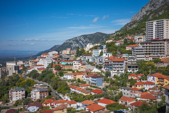 A stunning panoramic view of a European hillside city filled with red tiled rooftops, colorful houses, and modern apartment buildings rising toward dramatic rocky mountains