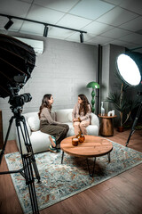 Two women converse on a modern indoor studio set with cameras, lights and microphones, filming an interview for broadcast or streaming